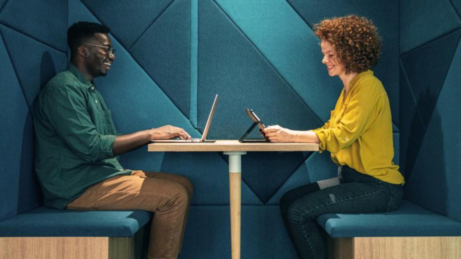 A man and a woman site opposite each other in a booth while working in a chic co-working office.