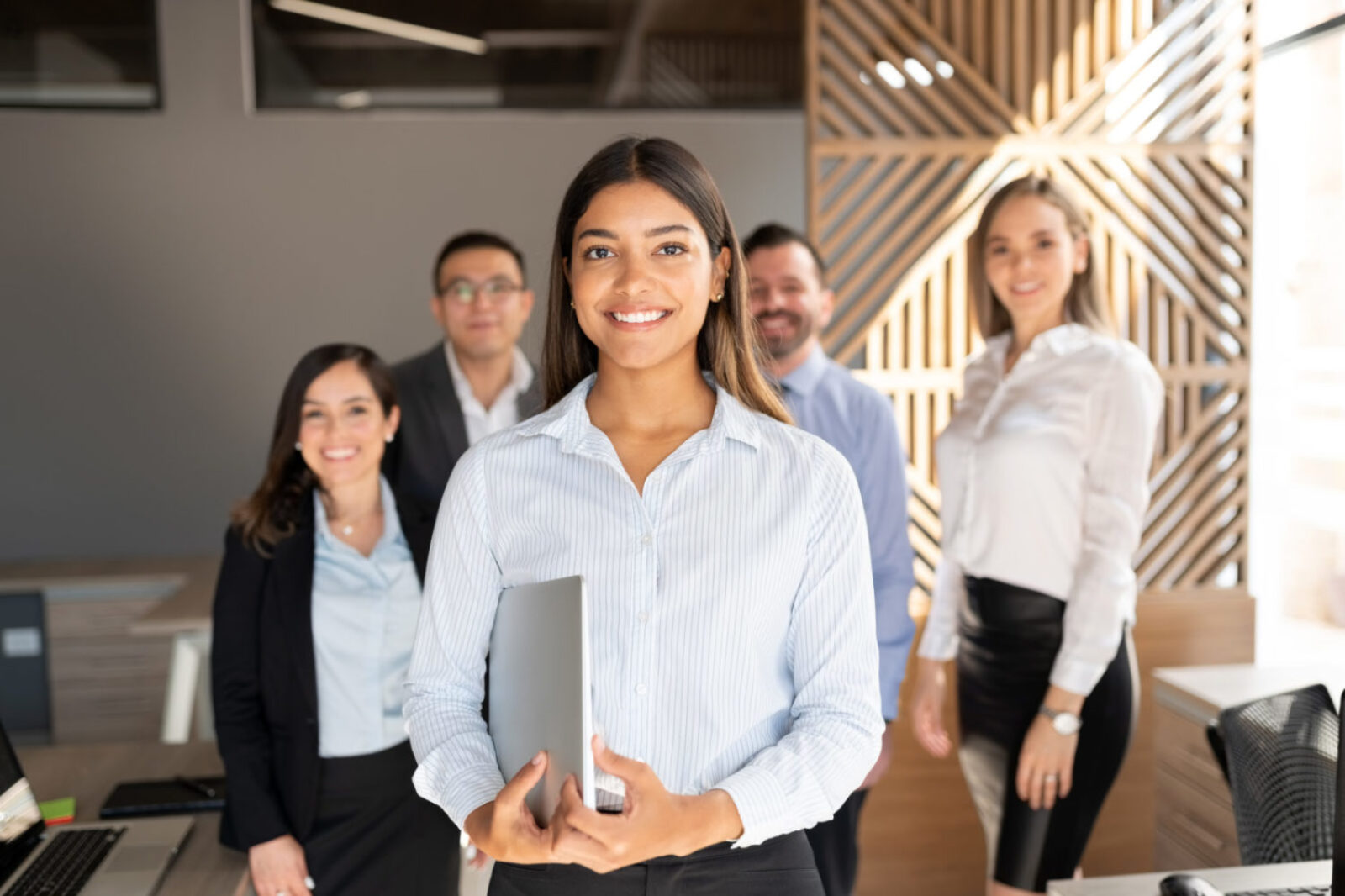 Confident hispanic businesswoman standing in office with colleagues in background